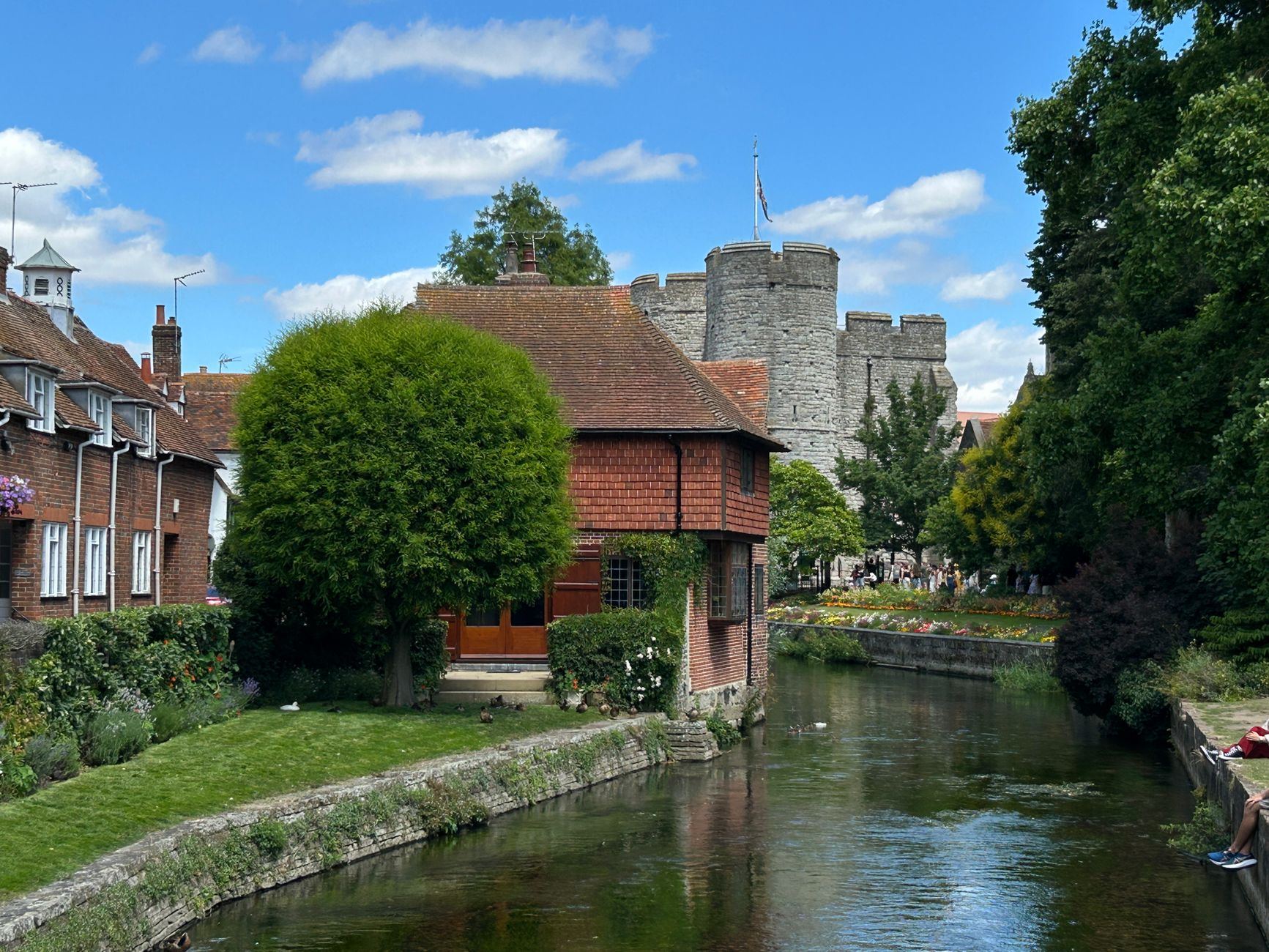 Picturesque English town scene with a castle, river, and charming houses under a bright summer sky.