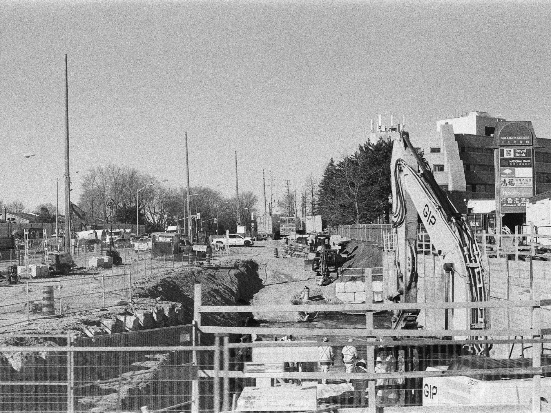 An excavator at a construction site with a trench and commercial buildings nearby