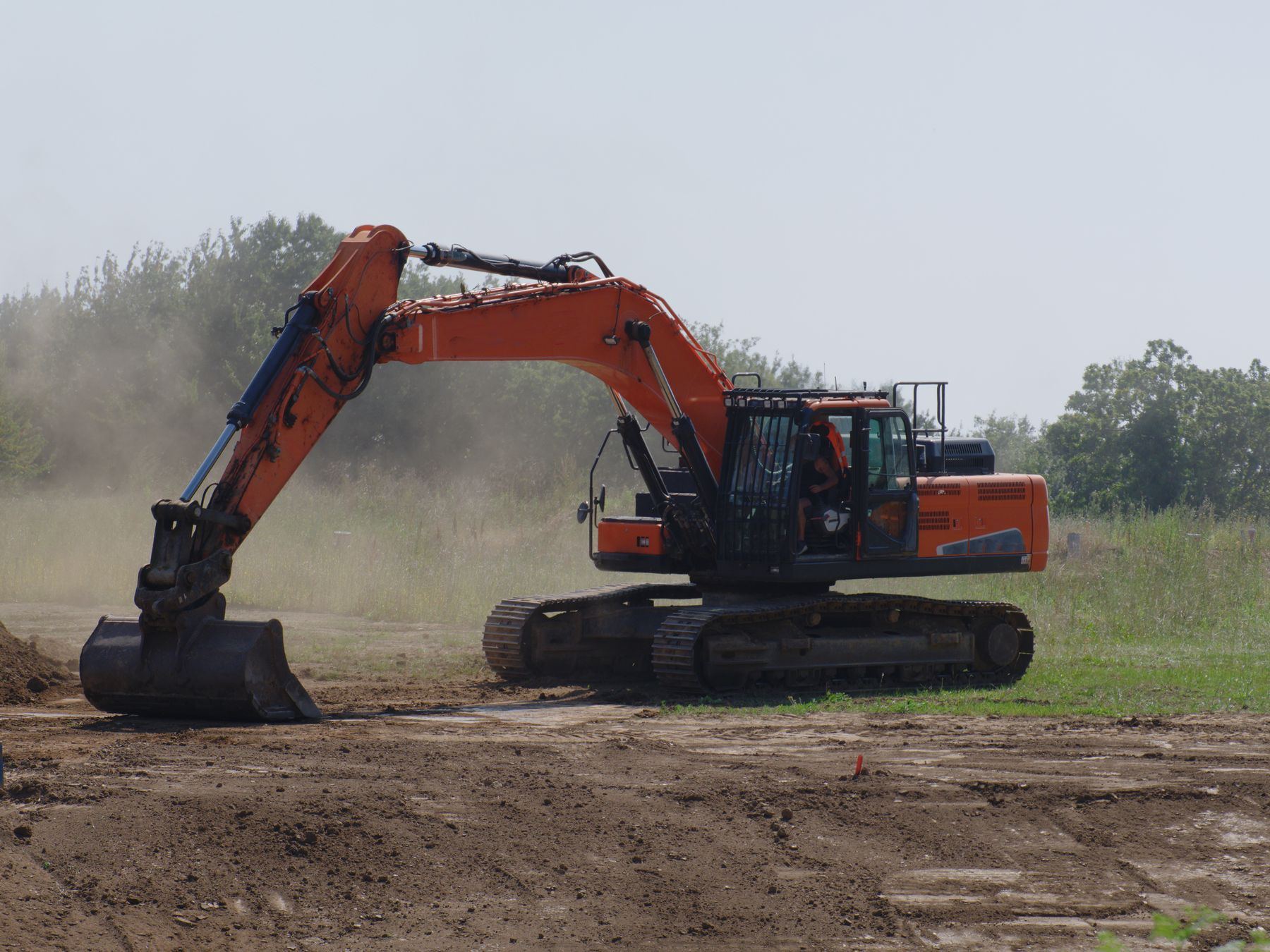 An excavator levels dirt at an outdoor construction site with dust and trees visible.
